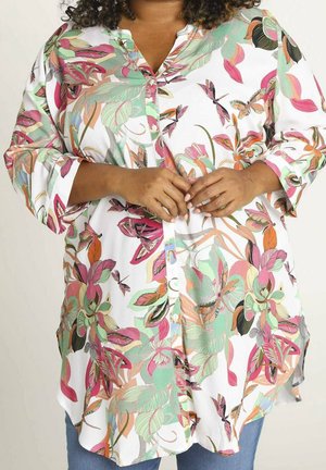 Plus-size persoon draagt een wit blouse met knopen en kleurrijke bloemen- en vlindermotieven, handen lichtjes voor elkaar gevouwen.
