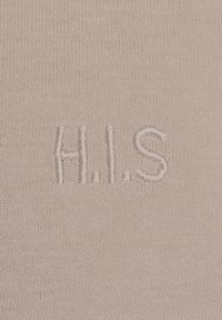 Lichtbeige stof met een gladde textuur, voorzien van geborduurde letters "H.I.S" in een iets donkerdere draad, gecentreerd op het materiaal.