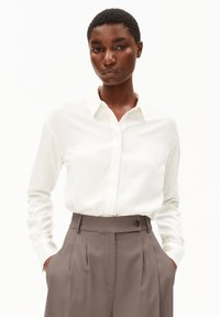 Camicia bianca a maniche lunghe con colletto, abbinata a pantaloni grigi a vita alta con dettaglio dei bottoni e tasche laterali. Tessuto liscio.
