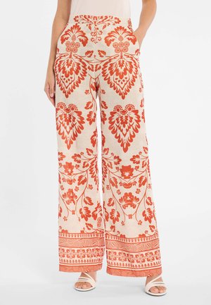 Beige Hosen mit weitem Bein, verziert mit kräftigen roten Blumen- und aufwändigen Mustern, Seitentaschen, kombiniert mit weißen Riemchensandalen und einem weißen Oberteil.