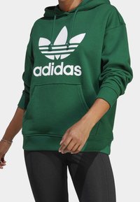 Grön hoodie i bomullsblandning, med en stor vit Adidas-logotyp och tre ränder på bröstet, samt en framficka.
