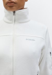 Persoană care poartă o jachetă polară albă cu fermoar frontal, guler ridicat și un mic logo Columbia brodat pe piept.