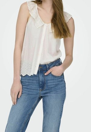Kvinde iført en ærmeløs hvid bluse med blonde detaljer og blå high-waisted jeans, den ene hånd i lommen og den anden hvilende ved siden.