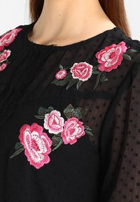 Blouse noire transparente avec des pois texturés et une broderie florale rose sur la poitrine et l'épaule, portée par une personne à la peau claire et aux cheveux longs.