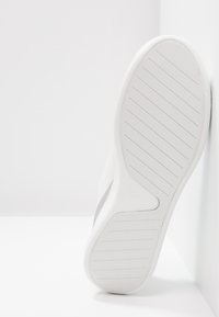 Zapatilla blanca con parte superior lisa y suela de goma texturizada. Presenta un diseño a rayas y un logo en el talón.