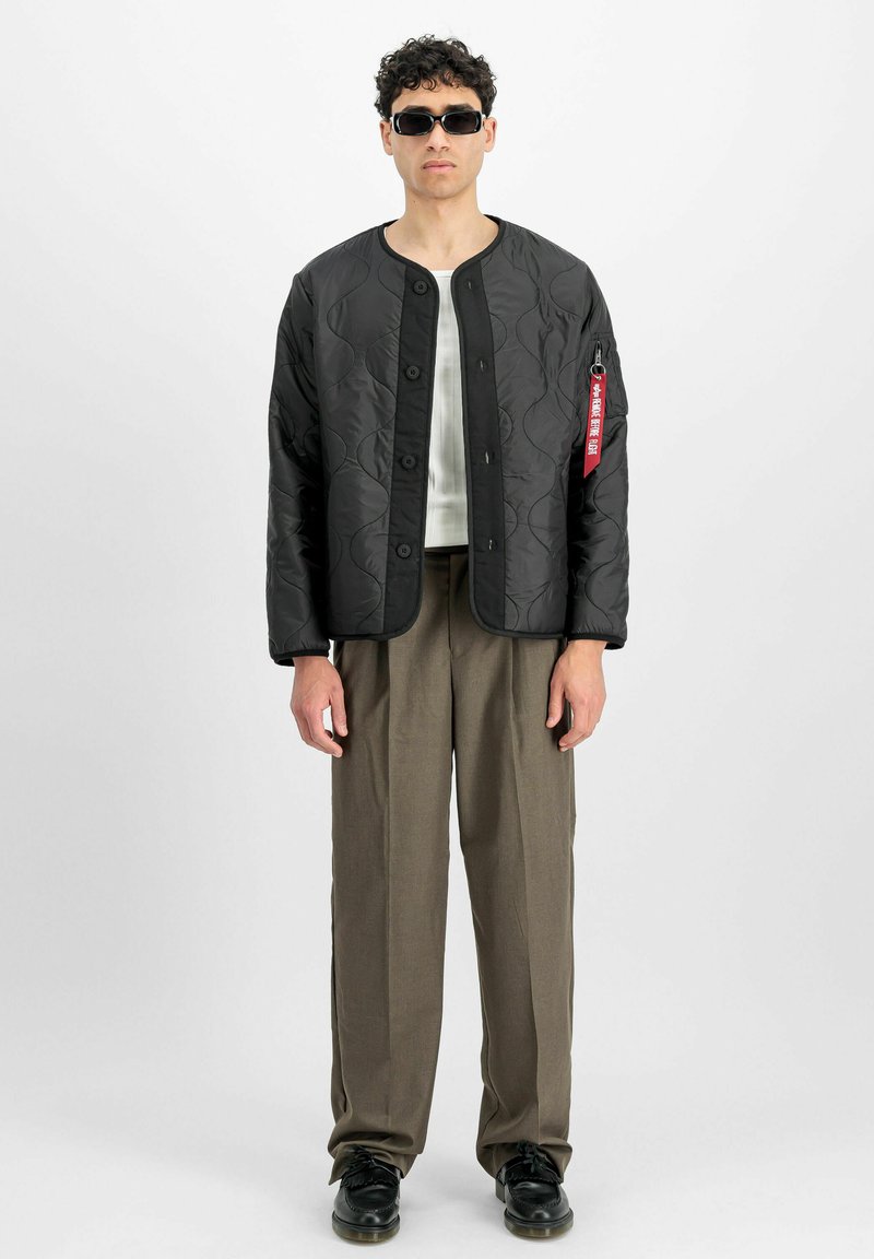 Veste matelassée noire avec un col arrondi, fermeture à boutons-pression, et un motif de vagues subtil, associée à un pantalon beige et des chaussures noires.