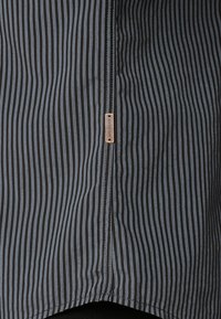 Gestreepte stof in marineblauw en lichtblauw, met verticale lijnen. Een klein, rechthoekig leren label is zichtbaar nabij de naad. Zachte textuur.