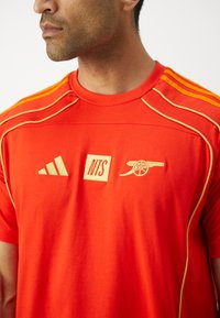 Rode sportieve t-shirt van katoen, met gele accenten, drie strepen op de schouders en geborduurde logo's: Adidas, NTS, Arsenal.