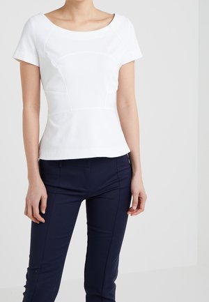 Kobieta w dopasowanym białym topie z krótkim rękawem i zakrzywionymi szwami oraz ciemnoniebieskich obcisłych spodniach, stojąca na jednolitym tle.