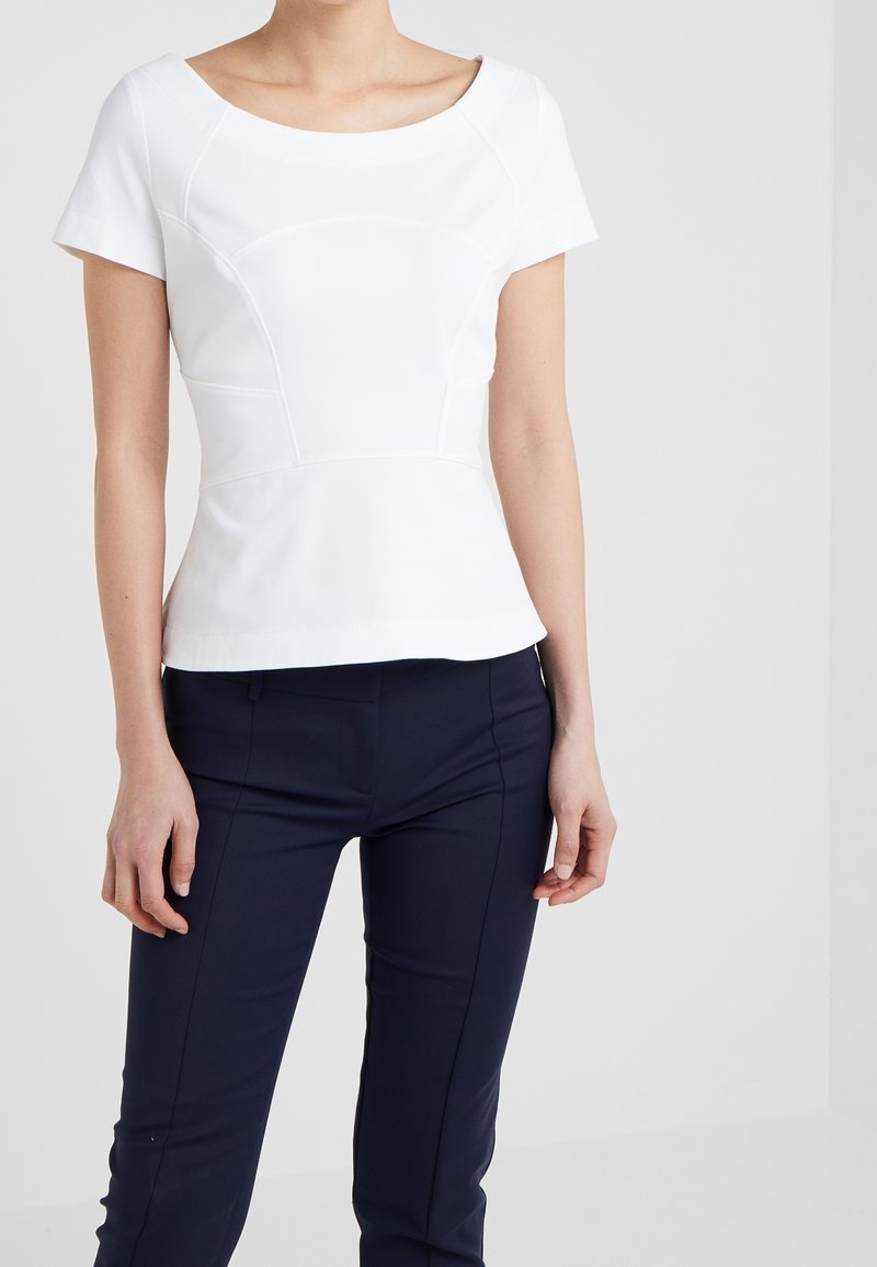 Femme portant un top blanc ajusté à manches courtes avec des détails de coutures courbes et un pantalon slim bleu marine foncé, debout devant un fond uni.