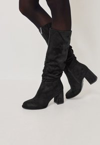 Schwarze knähöga stövlar i mjukt mocka, med en kraftig klack och en sidodragkedja. Designen är stilren med en något slapp passform.