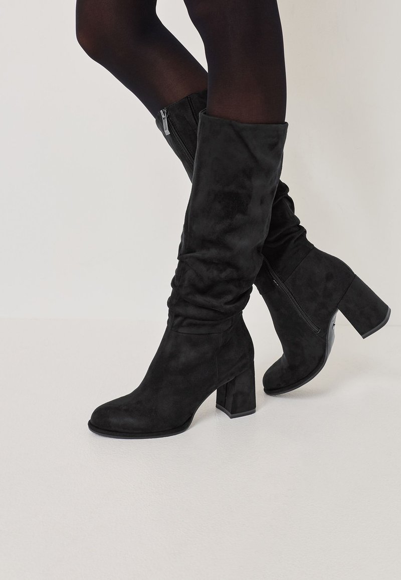 Schwarze knähöga stövlar i mjukt mocka, med en kraftig klack och en sidodragkedja. Designen är stilren med en något slapp passform.