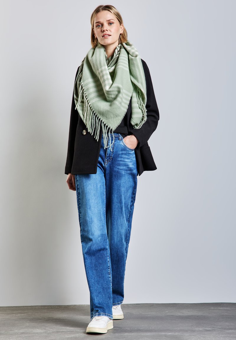 Écharpe vert menthe avec des franges, portée sur un manteau noir et un jean bleu. Le mannequin se tient sur une surface grise avec un arrière-plan neutre.