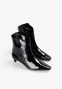 Botines de piel sintética negra con punta puntiaguda, tacón bajo y tachuelas decorativas en el lateral. Textura lisa y brillante en toda la superficie.