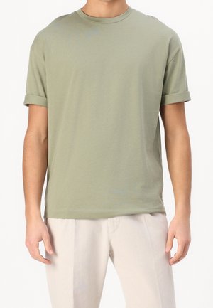 Man draagt lichtgroen kort mouw opgerolde T-shirt en lichtbeige broek, staand tegen een effen achtergrond.