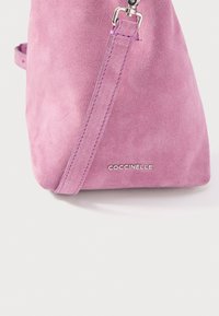 Ροζ τσάντα από σουέντ με ρυθμιζόμενο λουρί και ασημένιο μεταλλικό κούμπωμα, με λογότυπο «COCCINELLE» σε λείο, υφασμένο φόντο.
