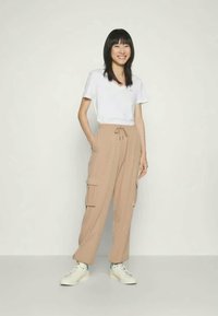 Usměvavá osoba stojící s rukama v kapsách béžových cargo kalhot, oblečená v bílém tričku a bílých teniskách na jednoduchém pozadí.