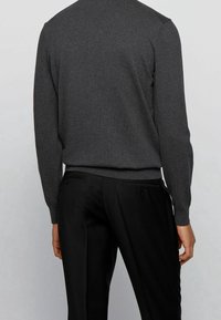 Person, die einen dunkelgrauen Langarmpullover und schwarze Hose trägt, von hinten vor einem einfarbigen hellgrauen Hintergrund.