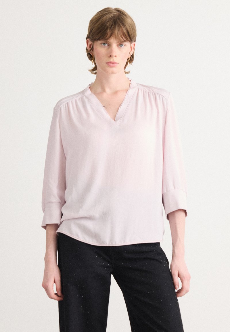 Blouse rose clair avec un décolleté en V et des manches trois-quarts. Présente un tissu doux, des fronces subtiles aux épaules et un ourlet arrondi.