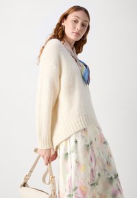 Suéter oversized color crema hecho de material suave y texturizado. Presenta puños y dobladillo acanalados. La modelo lleva una falda floral y sostiene un bolso beige.