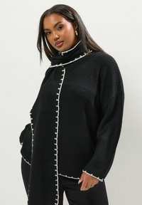Mustat neulepusero, jossa on korkea kaulus ja valkoiset kontrastipistoja reunoilla. Löysä malli pitkillä hihoilla ja teksturoitu rakenne.