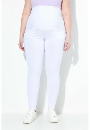 Kobieta w ciąży ubrana w białe legginsy ciążowe z wysokim stanem oraz białe trampki, stojąca na białym tle.