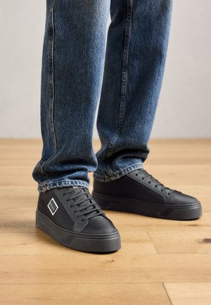 Person wearing dark blue jeans and black sneakers with a thick sole, standing on a light wooden floor.
