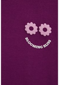 Camisa de algodón morada con dos apliques de flores rosas y texto blanco que dice "BLOOMING BLISS" en un diseño curvado en la parte inferior.