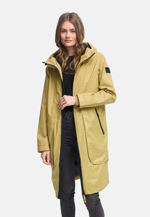 Jeune femme portant un long imperméable jaune à capuche par-dessus un haut noir et un jean foncé, debout devant un fond uni.