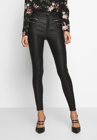 Pantalones ajustados de cuero sintético negro con bolsillos con cremallera en la parte frontal y detalles de costura, combinados con una blusa de manga larga con estampado floral y tacones negros.