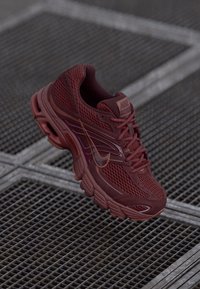 Bordová Nike teniska s sieťovinou a gumovou podrážkou, odpočívajúca na kovovej mriežke s geometrickým vzorom.