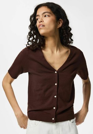 Femme aux cheveux bouclés portant un cardigan marron à manches courtes boutonné et un pantalon blanc, regardant vers sa gauche les mains sur les hanches.