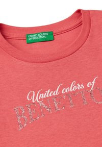 Coral T-shirt with a round neck. Features a green label and the text "United colors of BENETTON" in shimmering silver. Soft cotton texture.