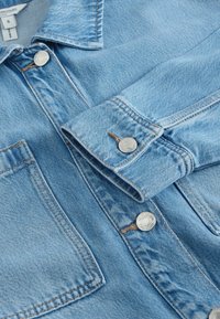 Veste en denim bleu clair avec boutons métalliques, col visible, poche poitrine et coutures piquées sur la manche repliée et le panneau avant.