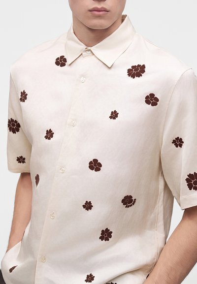 Chemise beige à manches courtes avec de petits motifs floraux marron foncé portée par une personne les mains dans les poches.