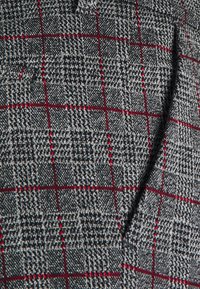 Tissu gris à motif à carreaux noir et rouge. Tissage texturé, montrant un détail de poche le long de la couture.