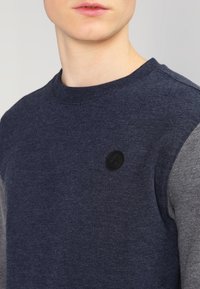 Granatowy sweatshirt z szarymi rękawami, z okrągłym dekoltem i czarnym okrągłym logo na piersi. Miękka tkanina z mieszanki bawełny.