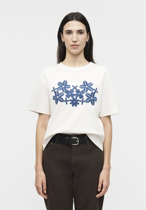 Γυναίκα με μακριά σκούρα μαλλιά φοράει λευκό κοντομάνικο πουκάμισο με μπλε κεντήματα λουλουδιών, μαύρη ζώνη και σκούρο παντελόνι, στέκεται μπροστά σε απλό φόντο.