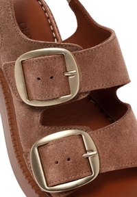 Sandal de ante marrón que presenta dos cintas anchas con grandes hebillas en tono dorado, una suela marrón duradera y un forro interior suave.