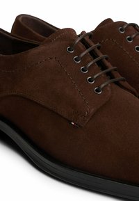 Zapatos de ante marrón con una textura suave, punta redonda y suela de goma negra. Presenta cordones marrones y detalles de costura. Etiqueta de acento tricolor visible.