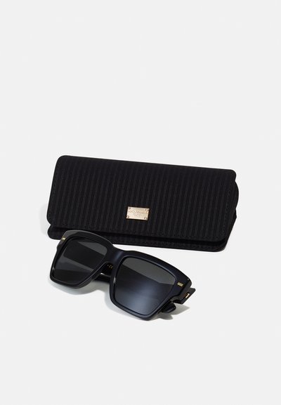 Black oversized sunglasses with dark lenses, square shape, and gold accents; accompanied by a striped black case with a metallic logo plate.