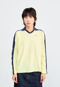 Chemise jaune à manches longues avec un motif à côtes verticales, col et manches bleu marine, dotée d'un petit logo doré sur la poitrine.