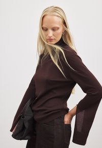 Brauner Langarmpullover mit hohem Kragen und glockigen Ärmeln. Glatte Textur, kombiniert mit dunklen Hosen und einer schwarzen Handtasche.