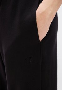 Μαύροι φόρμες από μαλακό ύφασμα, με πλαϊνή τσέπη και κεντητό μαύρο λογότυπο «cK». Ο σχεδιασμός είναι απλός και λιτός.