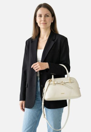 Vrouw draagt een zwart blazer en lichtblauwe jeans en houdt een witte handtas met gouden accenten vast tegen een effen achtergrond.
