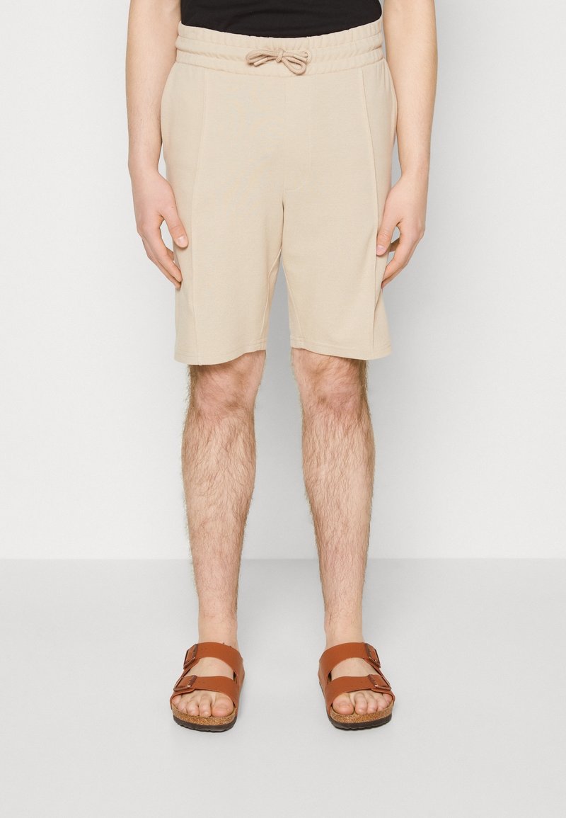 Homme portant un short beige mi-long à cordon et des sandales marron à double bride, debout devant un fond clair uni.