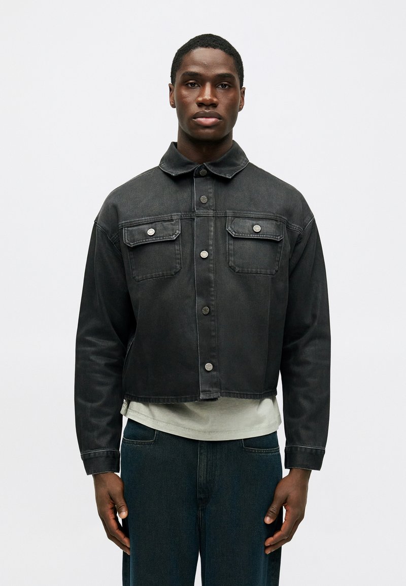 Črna denim jakna s skrajšanim dizajnom, koničastim ovratnikom, dvema žepoma na prsih in srebrnimi detajli. Nosite jo čez svetlo sivo srajco.