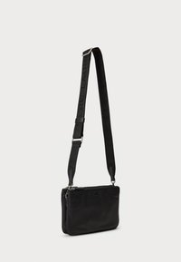 Sac bandoulière noir au design lisse et structuré, avec une sangle amovible ornée d'un logo embossé et une fermeture zippée.