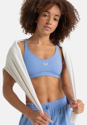 Light blue sports bra made of stretch fabric, featuring a small green logo at the center. White wrap draped over shoulders, matching blue shorts.