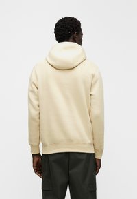 Bež pulover s kapuco z vrvico, rebrastimi manšetami in robom, izdelan iz mehke tkanine. Oblečen s temno zelenimi hlačami, viden od zadaj.